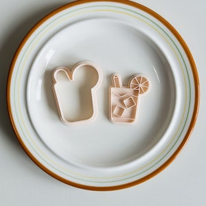 Pu&ograve; includere: Due stampini per biscotti in plastica su un piatto bianco con un bordo marrone. Gli stampini hanno la forma di un bicchiere di t&egrave; freddo con una fetta di limone e una fetta di pane.