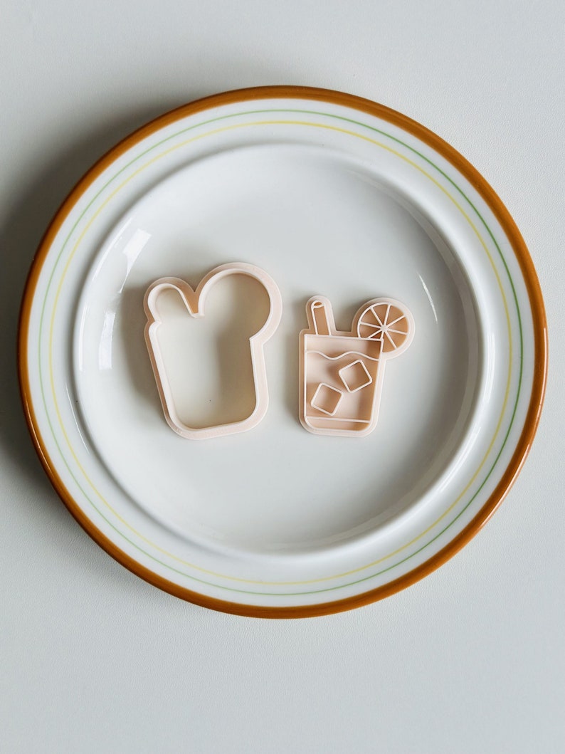 Pu&ograve; includere: Due stampini per biscotti in plastica su un piatto bianco con un bordo marrone. Gli stampini hanno la forma di un bicchiere di t&egrave; freddo con una fetta di limone e una fetta di pane.