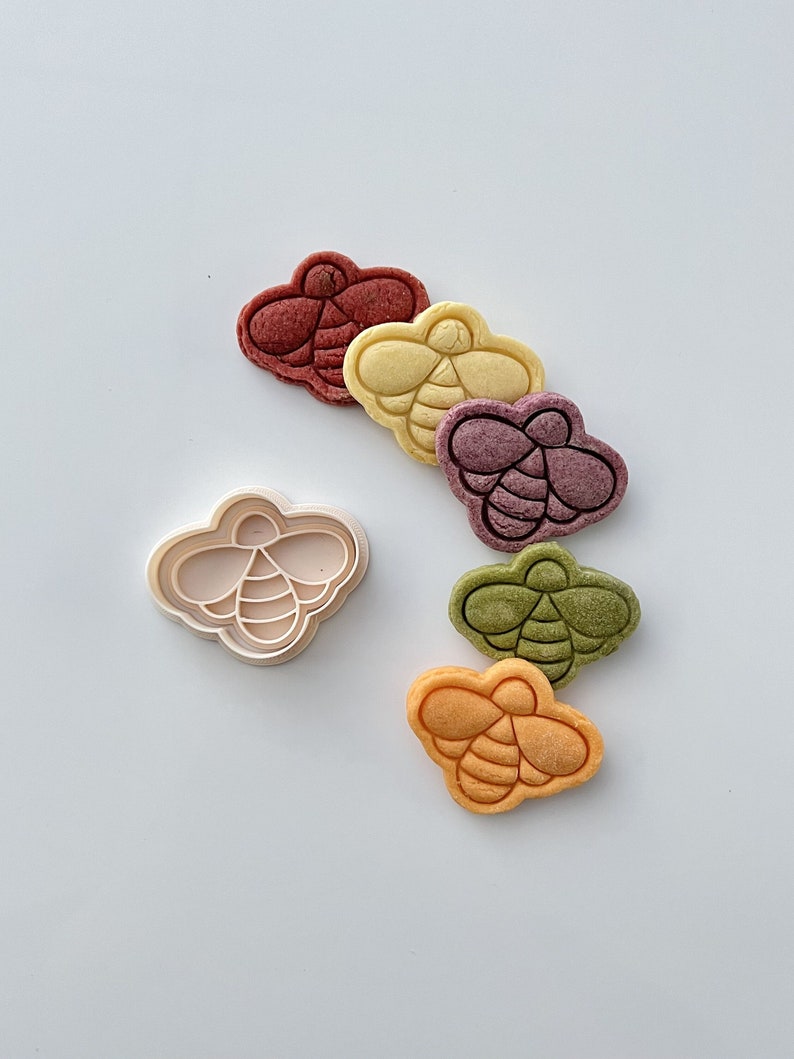May include: A white plastic bee-shaped cookie cutter with five colorful bee-shaped cookies. The cookies are red, yellow, purple, green, and orange.