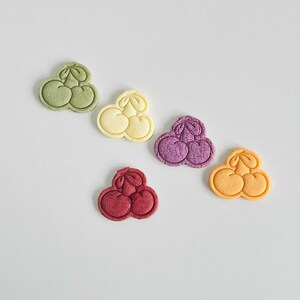 May include: Five colorful cookie cutters shaped like cherries. The cutters are green, yellow, purple, red, and orange.