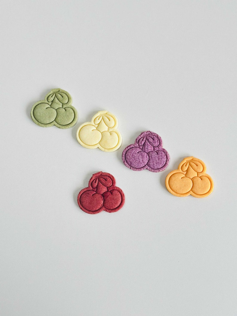 May include: Five colorful cookie cutters shaped like cherries. The cutters are green, yellow, purple, red, and orange.