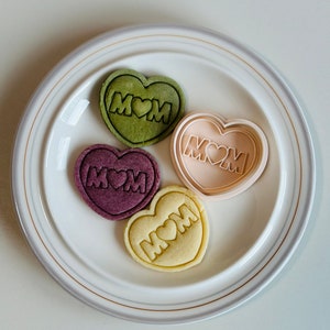 May include: Four heart-shaped cookies on a white plate with a gold rim. The cookies are decorated with the word "MOM" and a heart. The cookies are in shades of green, purple, beige, and yellow.