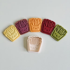 May include: Five colorful cookie cutters shaped like a carton of french fries with a smiling face. The cutters are arranged in a semi-circle with a white plastic cutter in the center.