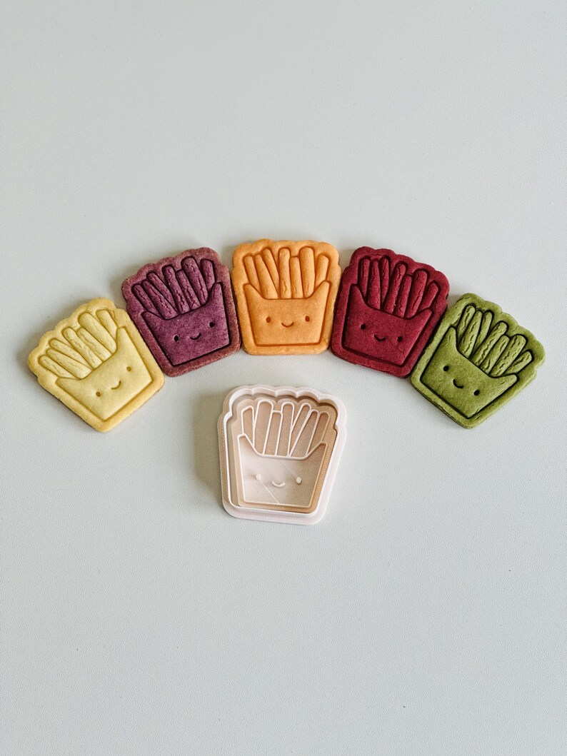 May include: Five colorful cookie cutters shaped like a carton of french fries with a smiling face. The cutters are arranged in a semi-circle with a white plastic cutter in the center.