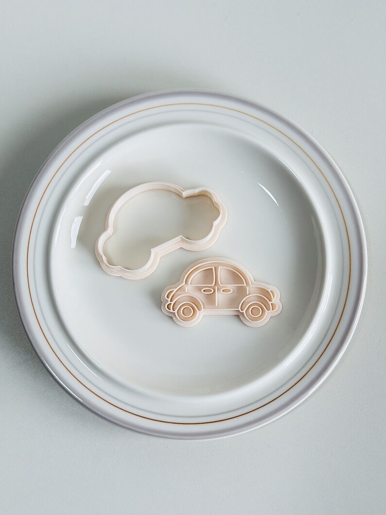 May include: Two plastic cookie cutters on a white plate with a gold rim. One cutter is shaped like a car and the other is shaped like a cloud.