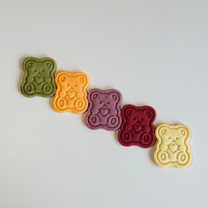 May include: Five bear-shaped cookies in a row, each a different color: green, yellow, purple, red, and white.
