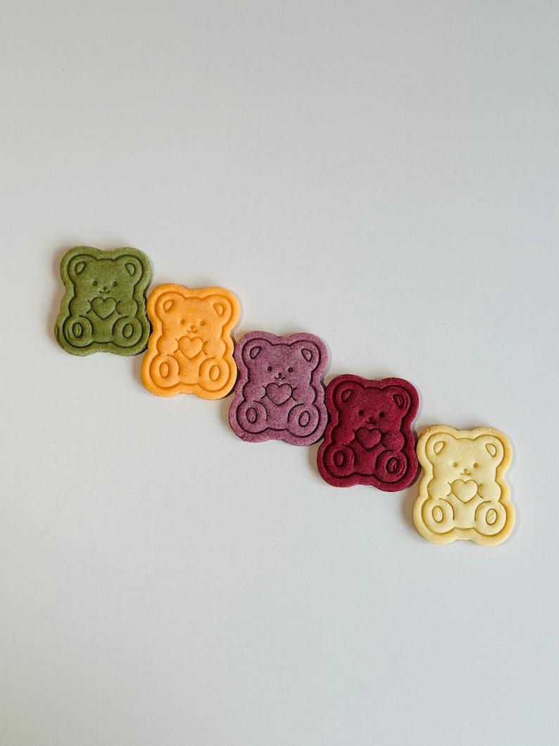 May include: Five bear-shaped cookies in a row, each a different color: green, yellow, purple, red, and white.
