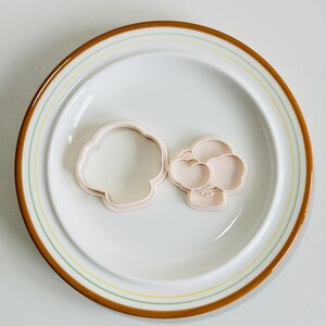 May include: Two plastic cookie cutters in the shape of a mushroom. The cutters are on a white plate with a brown and yellow rim.