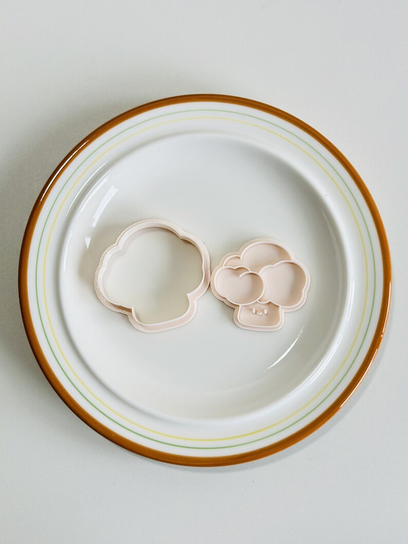 May include: Two plastic cookie cutters in the shape of a mushroom. The cutters are on a white plate with a brown and yellow rim.