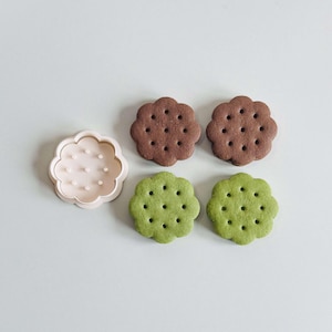 May include: Four flower-shaped cookies, two brown and two green, with a white plastic cookie cutter in the shape of a flower.