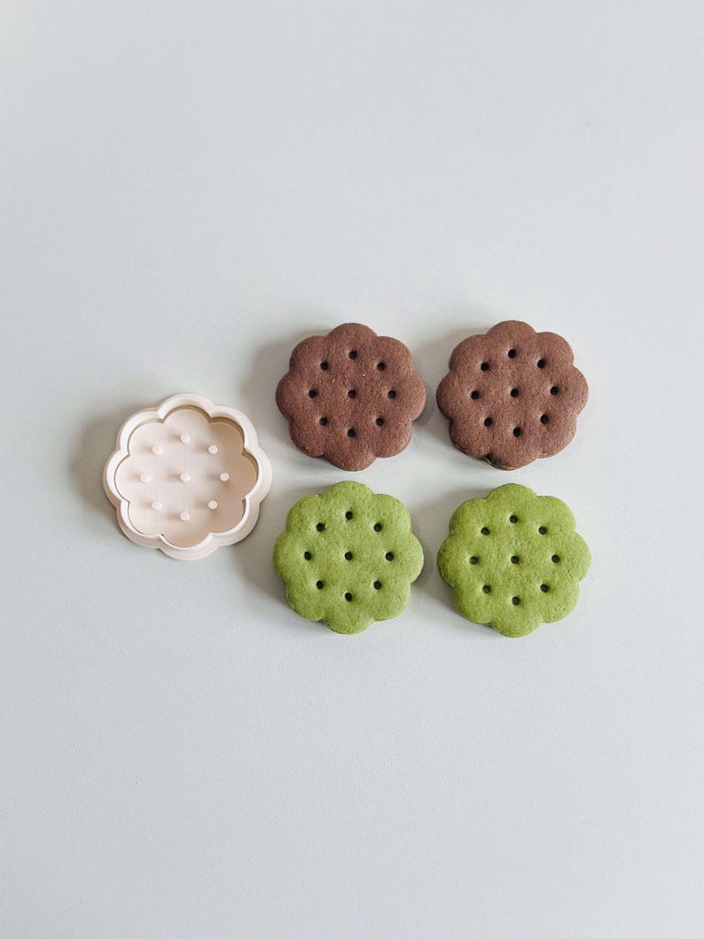 May include: Four flower-shaped cookies, two brown and two green, with a white plastic cookie cutter in the shape of a flower.