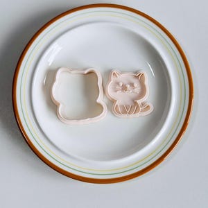 Puede incluir: Dos cortadores de galletas rosas con forma de gato sobre un plato blanco con rayas marrones y amarillas. Uno es un contorno, el otro un dise&ntilde;o detallado de gato. Ideal para hornear galletas con tem&aacute;tica de gatos.