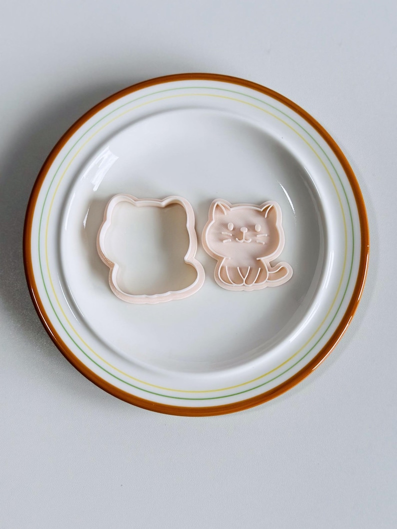 Puede incluir: Dos cortadores de galletas rosas con forma de gato sobre un plato blanco con rayas marrones y amarillas. Uno es un contorno, el otro un dise&ntilde;o detallado de gato. Ideal para hornear galletas con tem&aacute;tica de gatos.