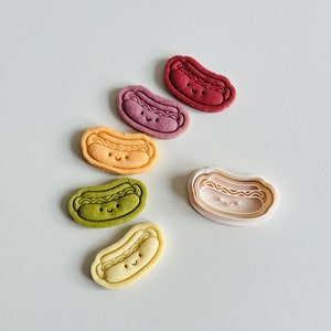 May include: Five colorful cookie cutters in the shape of a hot dog with a smiling face. The cutters are arranged in a semi-circle with the largest cutter in the center. The cutters are red, purple, orange, green, and yellow.