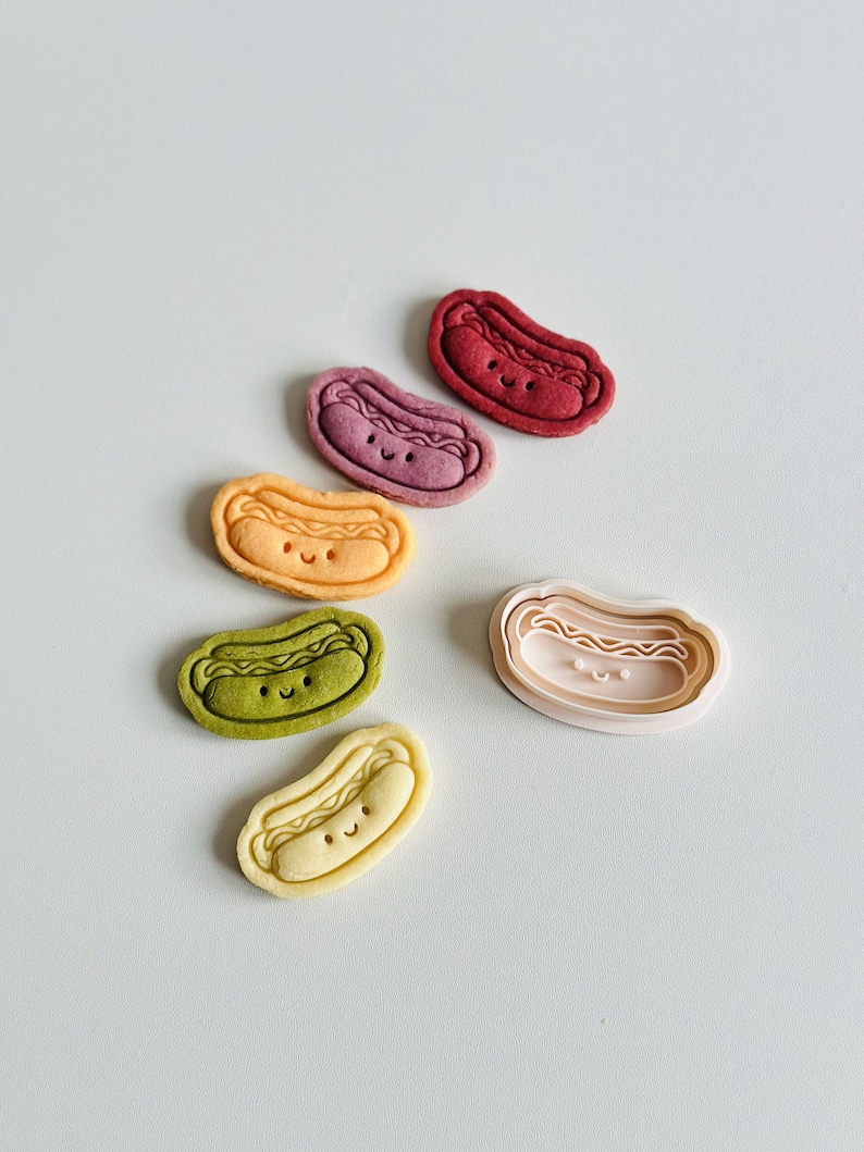 May include: Five colorful cookie cutters in the shape of a hot dog with a smiling face. The cutters are arranged in a semi-circle with the largest cutter in the center. The cutters are red, purple, orange, green, and yellow.