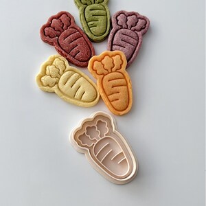 May include: Five carrot-shaped cookies in different colors, including green, purple, orange, and yellow, with a white plastic cookie cutter in the shape of a carrot.