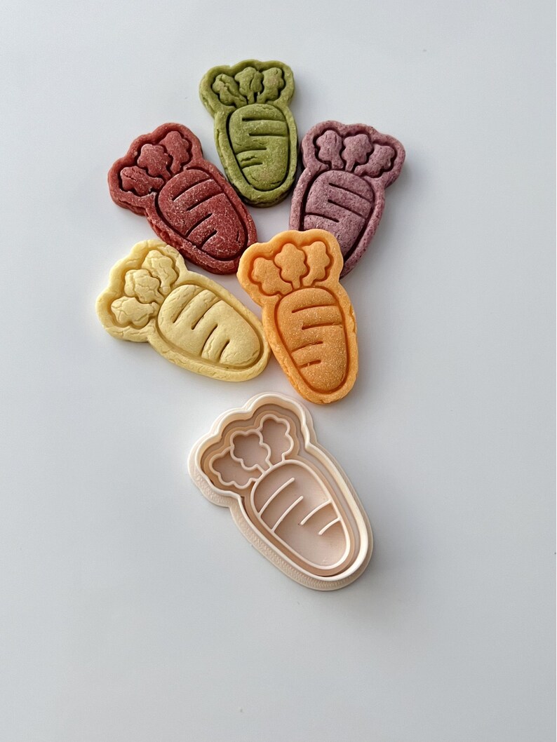 May include: Five carrot-shaped cookies in different colors, including green, purple, orange, and yellow, with a white plastic cookie cutter in the shape of a carrot.