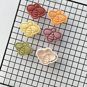 May include: Six bee-shaped cookies on a black wire cooling rack. The cookies are in various colors, including red, orange, yellow, green, and purple. A white plastic bee-shaped cookie cutter is also on the rack.