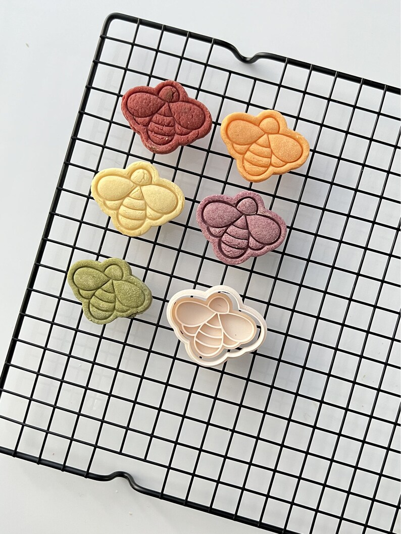 May include: Six bee-shaped cookies on a black wire cooling rack. The cookies are in various colors, including red, orange, yellow, green, and purple. A white plastic bee-shaped cookie cutter is also on the rack.