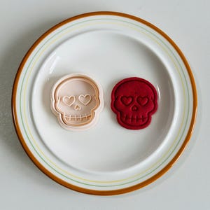 May include: Two skull-shaped items on a white plate with a brown and yellow rim. One is a light pink cookie cutter, and the other is a red skull-shaped item. Both have heart-shaped eye sockets and a row of teeth.