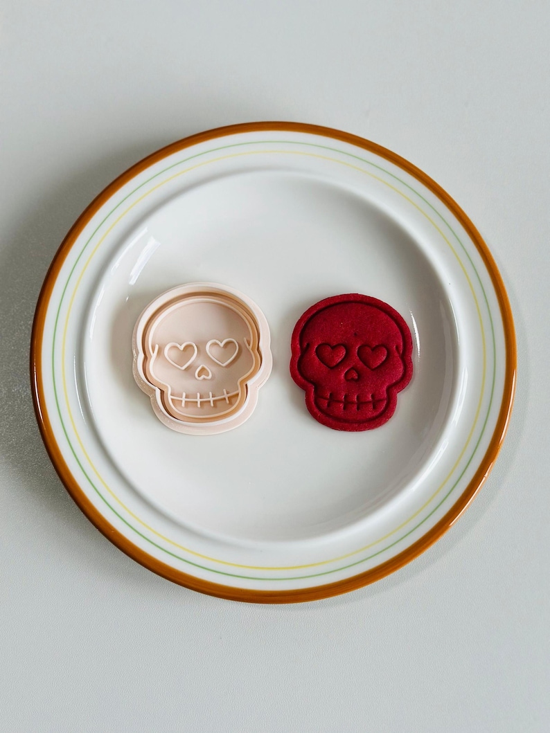 May include: Two skull-shaped items on a white plate with a brown and yellow rim. One is a light pink cookie cutter, and the other is a red skull-shaped item. Both have heart-shaped eye sockets and a row of teeth.