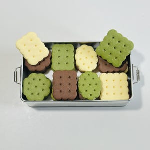 May include: A silver metal container filled with a variety of sandwich cookies. The cookies are green, brown, and yellow and have a patterned design.