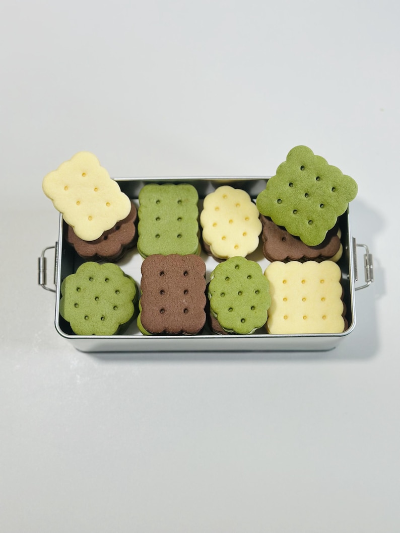 May include: A silver metal container filled with a variety of sandwich cookies. The cookies are green, brown, and yellow and have a patterned design.