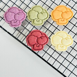 May include: Five colorful broccoli-shaped cookies on a black wire rack. The cookies are decorated with a simple smiley face and are in shades of purple, green, orange, red, and yellow.