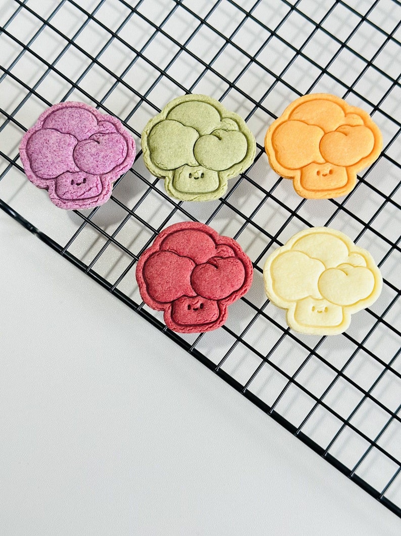 May include: Five colorful broccoli-shaped cookies on a black wire rack. The cookies are decorated with a simple smiley face and are in shades of purple, green, orange, red, and yellow.
