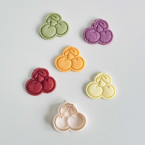 May include: Five cookie cutters in the shape of three cherries, in various colors: green, purple, orange, red, and white. The white cutter is the only one that is not a cookie.