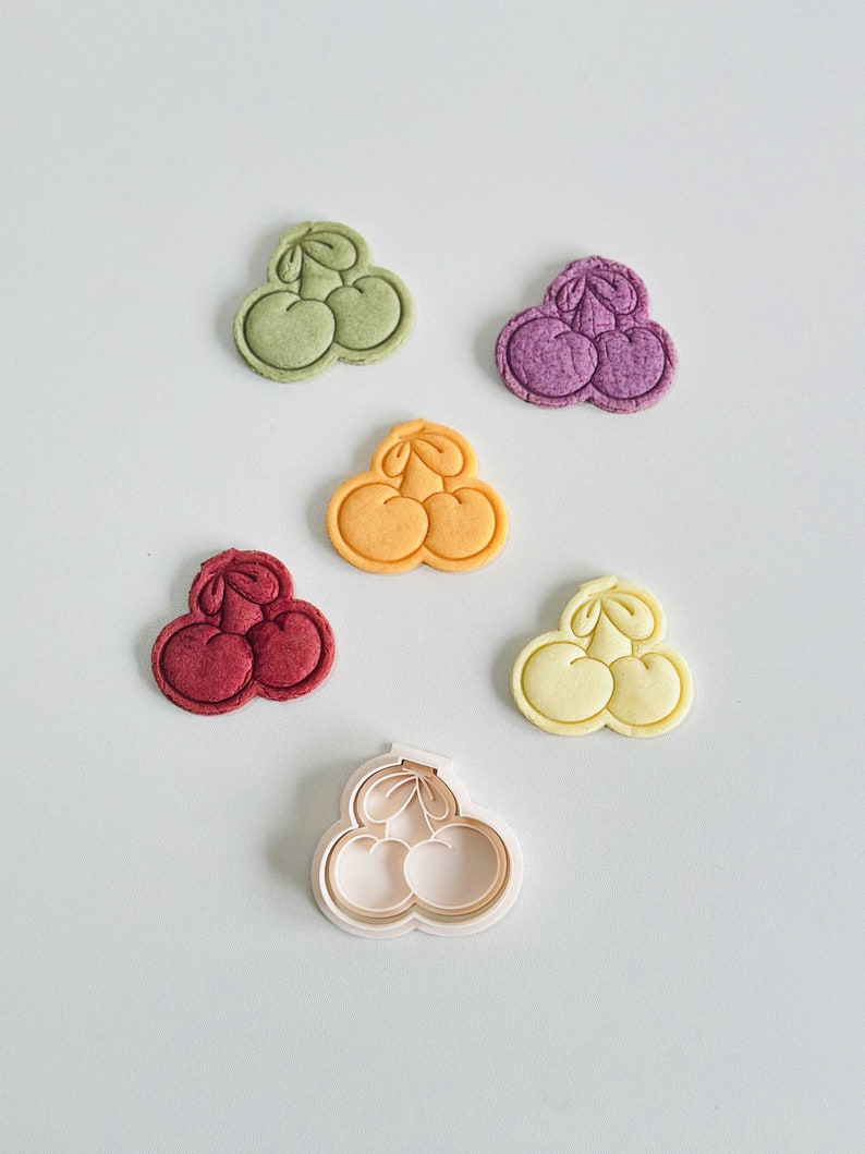 May include: Five cookie cutters in the shape of three cherries, in various colors: green, purple, orange, red, and white. The white cutter is the only one that is not a cookie.