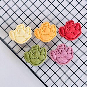 Op de afbeelding: Zes vogelvormige koekjes in verschillende kleuren, waaronder geel, oranje, rood, groen en roze, gerangschikt op een zwart draadkoelrek. Elk koekje heeft een gedetailleerd vogelontwerp met vleugels en een snavel.