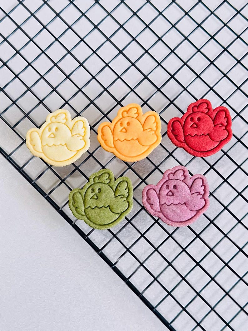 Op de afbeelding: Zes vogelvormige koekjes in verschillende kleuren, waaronder geel, oranje, rood, groen en roze, gerangschikt op een zwart draadkoelrek. Elk koekje heeft een gedetailleerd vogelontwerp met vleugels en een snavel.