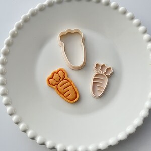 May include: Three cookie cutters on a white plate with a beaded edge. The cutters are shaped like carrots and a single carrot cookie is on the plate.