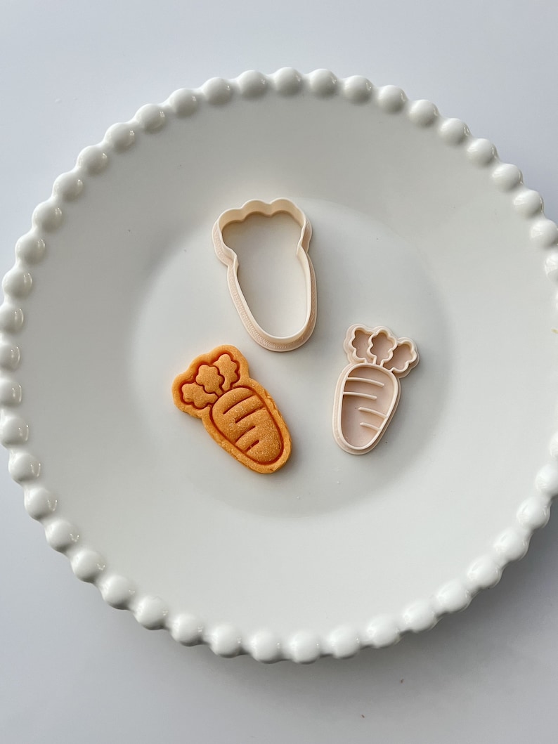 May include: Three cookie cutters on a white plate with a beaded edge. The cutters are shaped like carrots and a single carrot cookie is on the plate.
