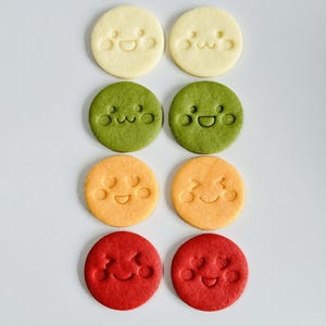 May include: Eight round cookies in a grid, each with a smiling face design. The cookies are in four colors: yellow, green, orange, and red. The cookies are arranged on a white surface.