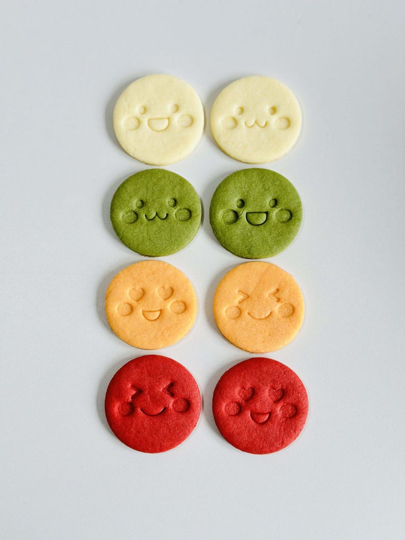 May include: Eight round cookies in a grid, each with a smiling face design. The cookies are in four colors: yellow, green, orange, and red. The cookies are arranged on a white surface.