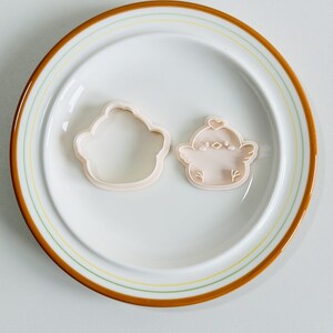 Op de afbeelding: Twee lichtroze koekjessnijders in de vorm van een cupcake en een kuiken, op een witte plaat met een bruine en gele rand. De kuikensnijder heeft een hartje op zijn hoofd en kleine vleugeld details. De plaat heeft een diameter van ongeveer 20 cm.