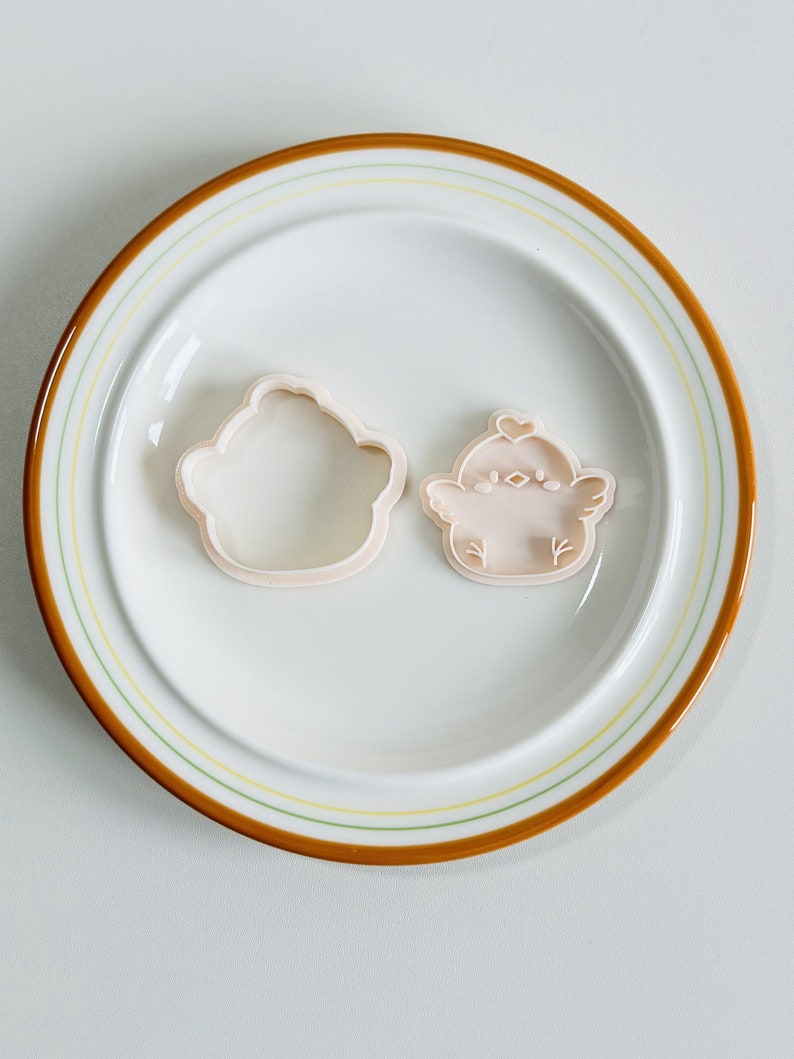 Op de afbeelding: Twee lichtroze koekjessnijders in de vorm van een cupcake en een kuiken, op een witte plaat met een bruine en gele rand. De kuikensnijder heeft een hartje op zijn hoofd en kleine vleugeld details. De plaat heeft een diameter van ongeveer 20 cm.