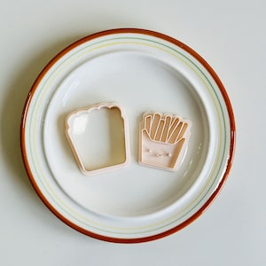 May include: Two pink plastic cookie cutters on a white plate with a brown and yellow rim. One cutter is shaped like a loaf of bread and the other is shaped like a container of french fries.