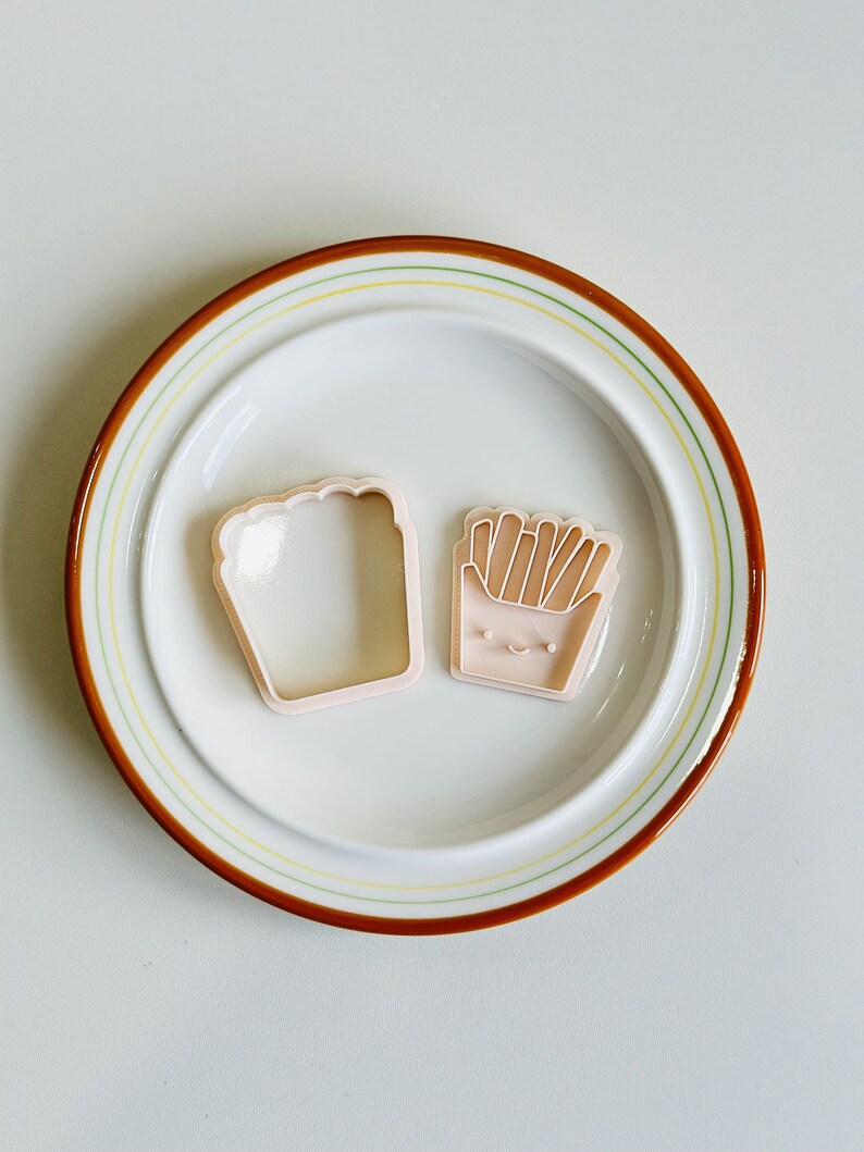 May include: Two pink plastic cookie cutters on a white plate with a brown and yellow rim. One cutter is shaped like a loaf of bread and the other is shaped like a container of french fries.