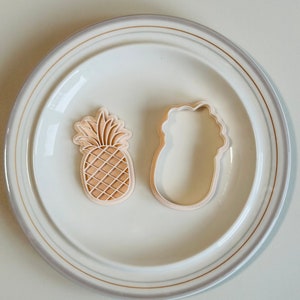 May include: Two cookie cutters, one shaped like a pineapple and the other shaped like a flower, on a white plate with a gold rim.