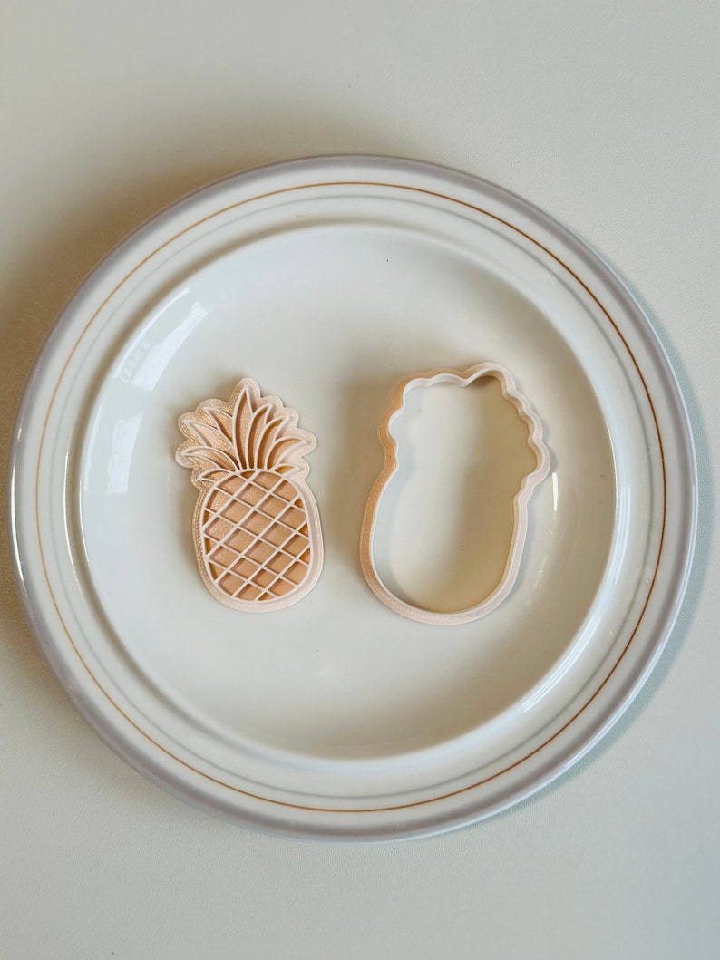 May include: Two cookie cutters, one shaped like a pineapple and the other shaped like a flower, on a white plate with a gold rim.