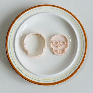 May include: Two light pink skull-shaped cookie cutters on a white plate with a brown and yellow rim. One cutter is a simple outline, the other has heart-shaped eyes and stitched mouth detail. Ideal for baking and Halloween.