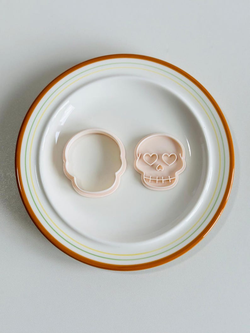 May include: Two light pink skull-shaped cookie cutters on a white plate with a brown and yellow rim. One cutter is a simple outline, the other has heart-shaped eyes and stitched mouth detail. Ideal for baking and Halloween.