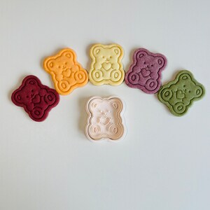 May include: Five bear-shaped cookies in different colors, including red, purple, green, yellow, and orange, with a white plastic bear-shaped cookie cutter in the center.