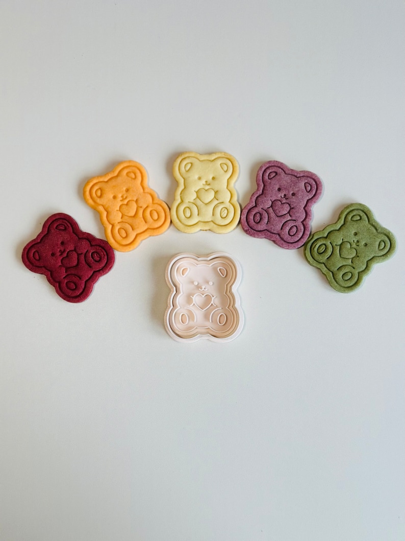 May include: Five bear-shaped cookies in different colors, including red, purple, green, yellow, and orange, with a white plastic bear-shaped cookie cutter in the center.