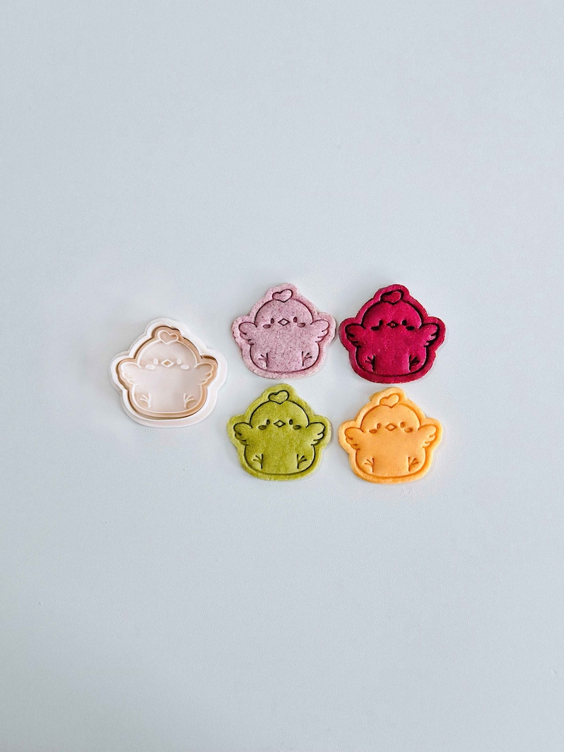 Op de afbeelding: Zes items in de vorm van een kuiken worden getoond. E&eacute;n is een lichtroze koekjesvorm, en de andere zijn koekjes in roze, rood, groen en oranje. Elk kuiken heeft een hart op zijn hoofd en kleine vleugels.