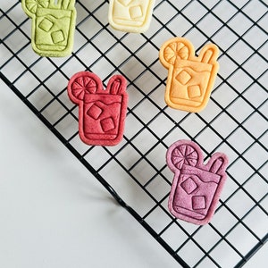 Pu&ograve; includere: Cinque biscotti a forma di bevanda ghiacciata su una griglia di raffreddamento in filo metallico nero. I biscotti hanno la forma di un bicchiere con una fetta di limone e una cannuccia. I biscotti sono gialli, arancioni, verdi, rossi e viola.