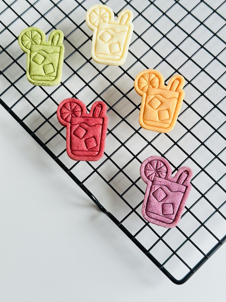 Pu&ograve; includere: Cinque biscotti a forma di bevanda ghiacciata su una griglia di raffreddamento in filo metallico nero. I biscotti hanno la forma di un bicchiere con una fetta di limone e una cannuccia. I biscotti sono gialli, arancioni, verdi, rossi e viola.